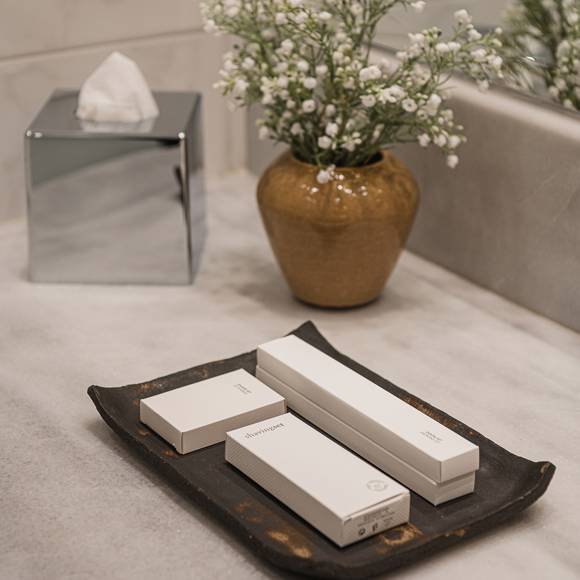 Bathroom amenities on a ceramic tray, beside a vase with white flowers on marble surface