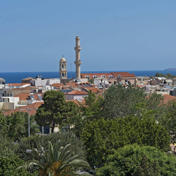 Old Town rooftops with a minaret and church bell tower, seen above green treetops and the sea
