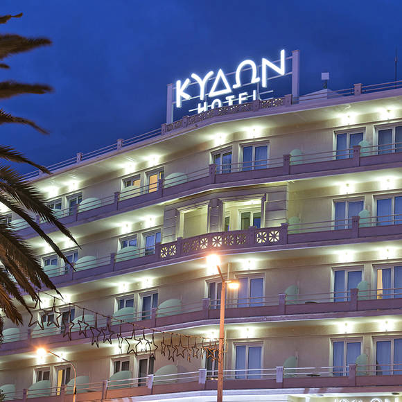 Illuminated hotel façade with lit balconies and rooftop sign at dusk, framed by a palm tree